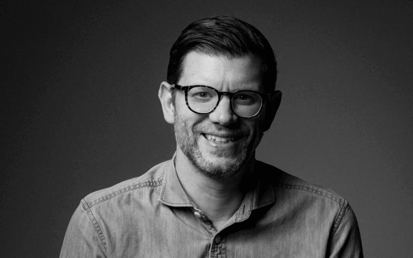 A black-and-white portrait of a person wearing glasses and a button-up shirt, smiling against a plain background.