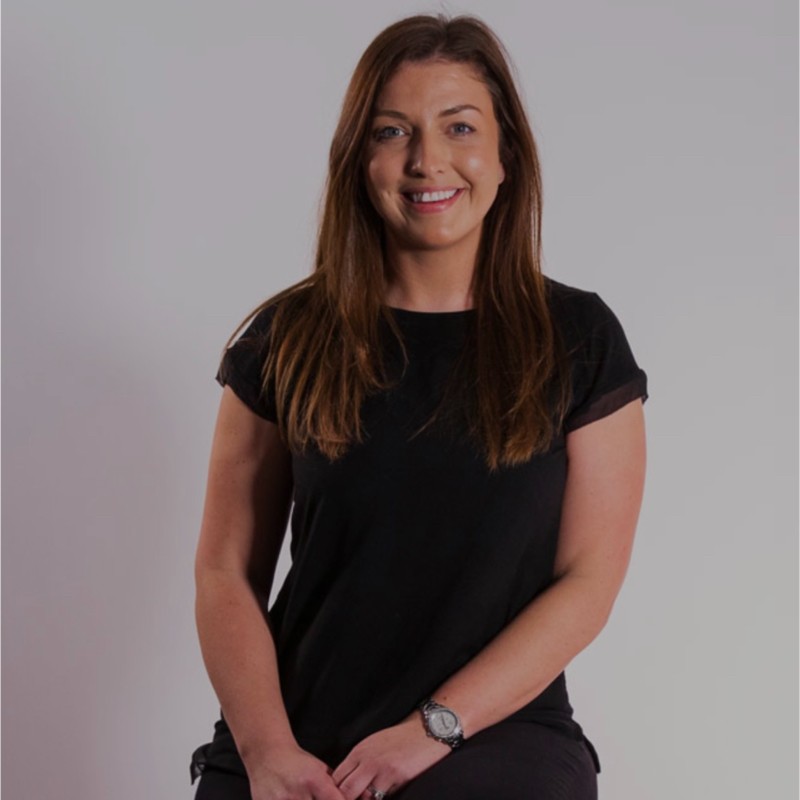 Woman sitting against a neutral background, wearing a black shirt, smiling with hands clasped in front.