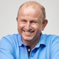 A man smiling warmly, wearing a blue collared shirt, against a plain background.