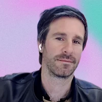 Man with dark hair and a beard wearing a leather jacket and wireless earbuds, set against a pastel gradient background.