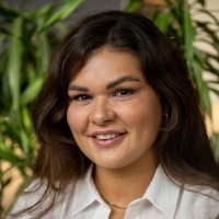 A woman with dark hair smiles warmly, wearing a white shirt, with green plants in the background.