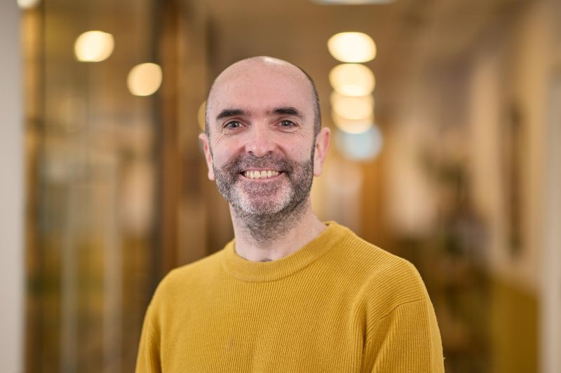 A smiling man in a mustard sweater stands in a warmly lit hallway, creating a friendly and approachable atmosphere.
