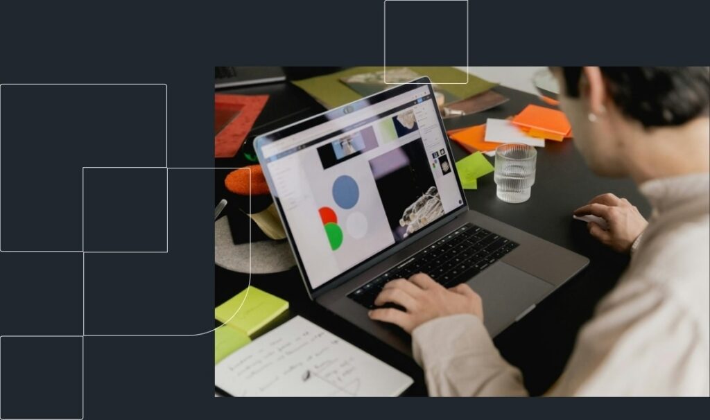 A person working on a laptop displaying colorful charts. The desk is cluttered with papers, post-it notes, and a clear glass of water.