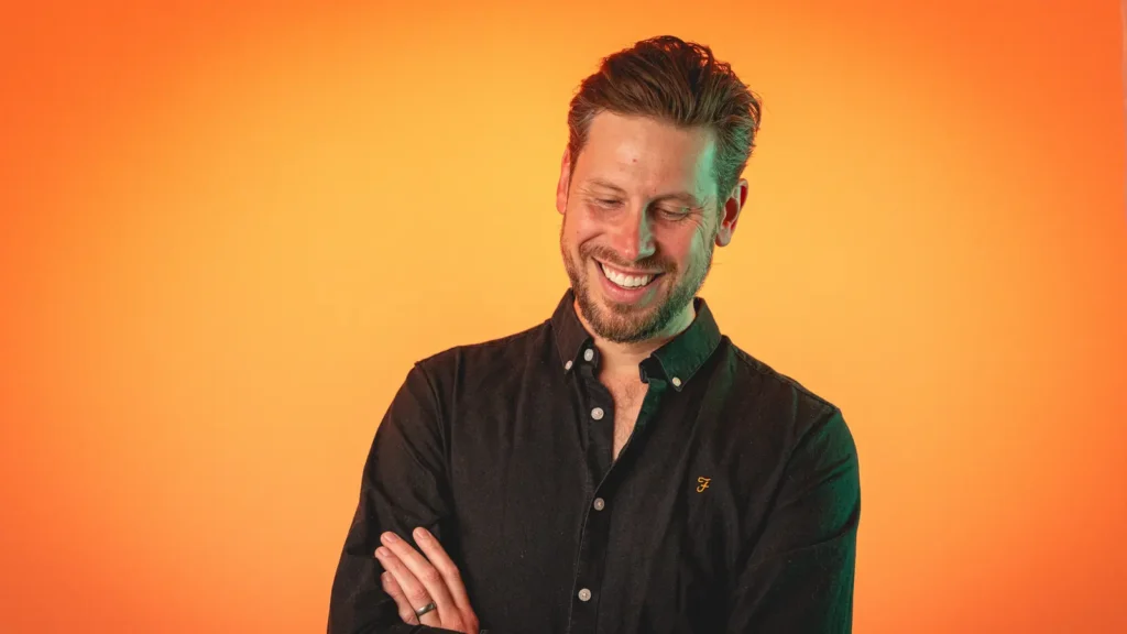 Man in a black shirt smiling with arms crossed against an orange background.