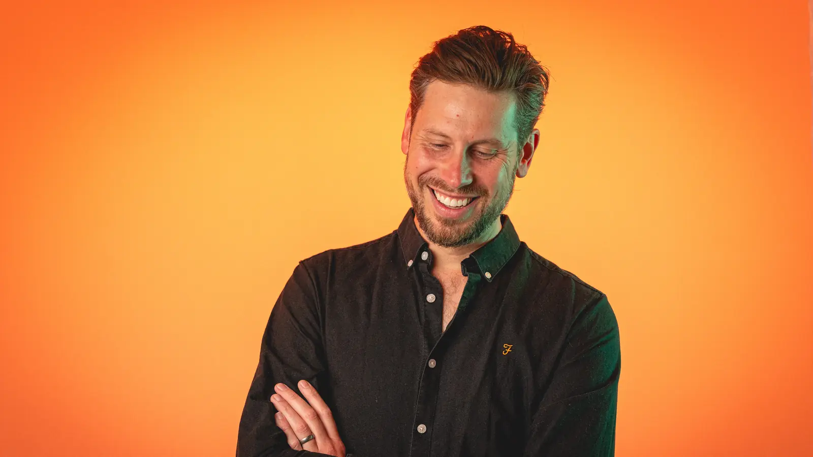 Man in a black shirt smiling with arms crossed against an orange background.