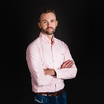 Man in a pink shirt and jeans stands confidently against a black background, arms crossed.