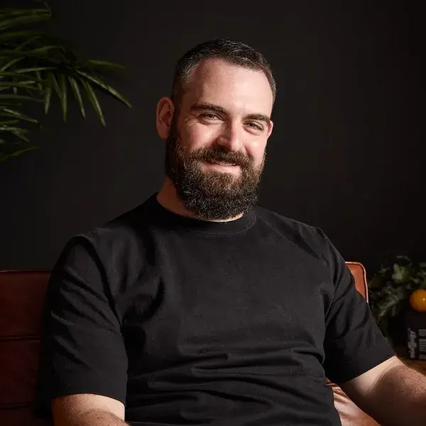 Bearded man in a black t-shirt, sitting on a brown leather couch with plants in the background.