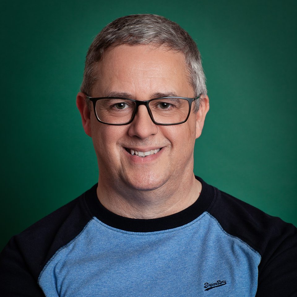 A smiling man with short gray hair and glasses wearing a blue and black sweatshirt against a green background.